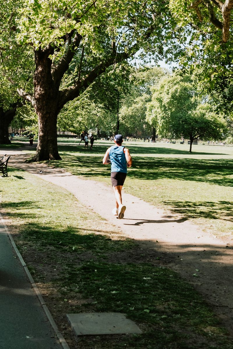 L'ostéopathie accompagne le coureur, de la prévention à la récupération. Coureur en camisole bleue sur un sentier de parc ombragé - Clinique Synergek
